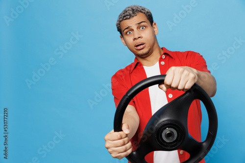 Young shcoked sad man of African American ethnicity 20s he wear red shirt hold steering wheel pretend driving look aside isolated on plain pastel light blue cyan background People lifestyle concept