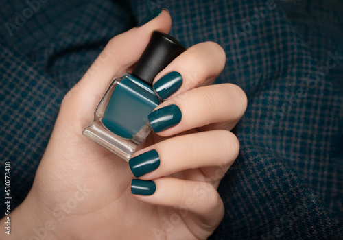 Female hand with dark nail design. Glitter turquoise nail polish manicure. Woman hand hold turquoise nail polish bottle.