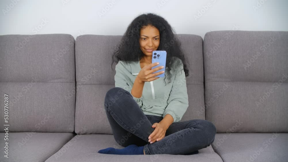 Young black woman using smartphone. African female person sitting on sofa and browsing modern mobile phone with triple camera. BIPOC person lifestyle video clip Young black woman using smartphone. African female person sitting on sofa and browsing modern mobile phone with triple camera. BIPOC person lifestyle video clip