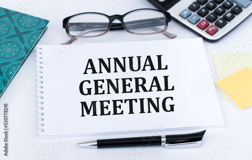 AGM Annual General Meeting is written in red on a white piece of paper on a light yellow background next to a laptop, pen, magnifying glass, glasses and a green plant.