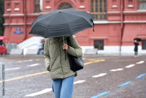Rain in city, slim girl in jeans and jacket walk with black umbrella on a street. Rainy weather in autumn