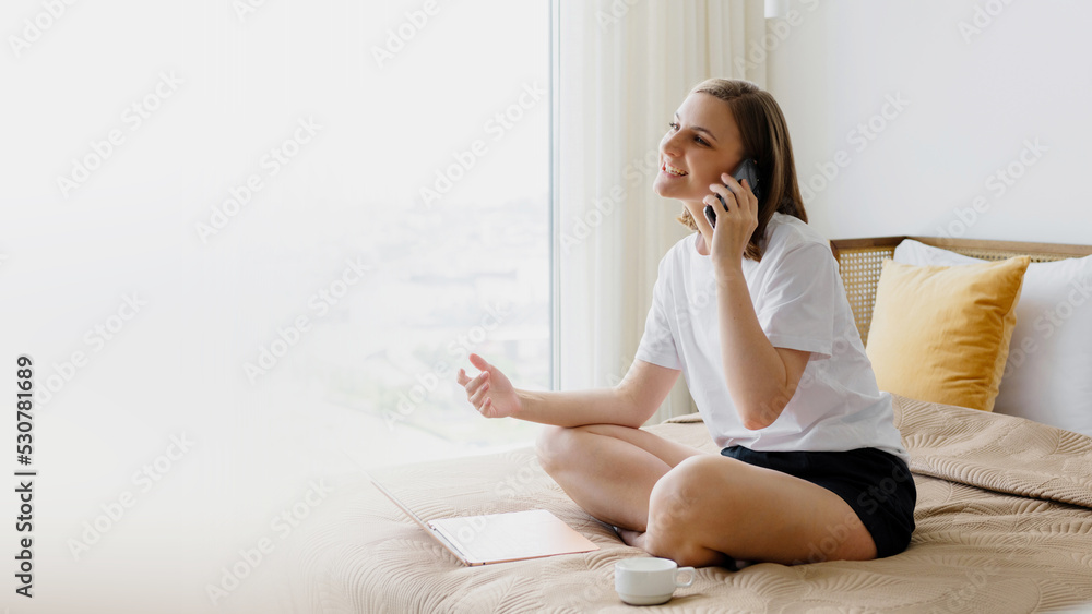 Young woman sitting on the bed in front of a laptop and talking on the phone actively gesturing with her hands Young woman sitting on the bed in front of a laptop and talking on the phone actively gesturing with her hands