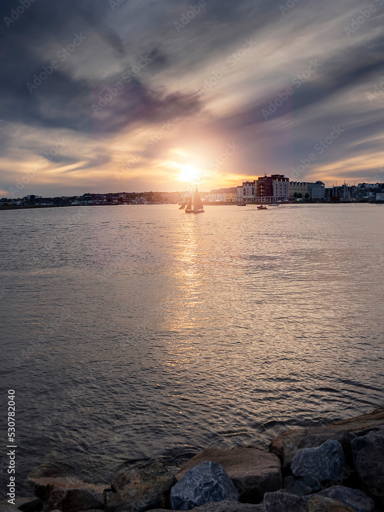 Naklejka premium Silhouette of Galway hooker type wooden boat sailing from harbor at stunning sunset time. Hobby and water sport. Dark and moody sky and dark water of Galway bay, Ireland.