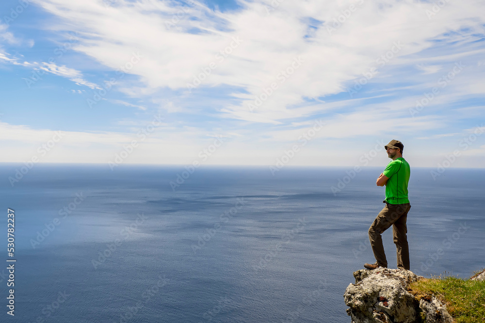 Man standing on an edge of a rock and looking at vast ocean and blue cloudy sky in the background. Reaching you goals and self belief concept. Copy space. Travel and tourism concept.