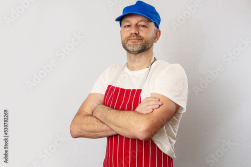 Portrait of professional butcher in white t shirt and red and white stripes classic apron and blue baseball cap. Meat industry and food supply. Male in 40s with grey beard and slim athletic body type.