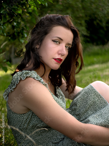 Outddor oniric portrait of a caucasian brunette  woman with red lipstick. She's wearing a green summer dress. Blurred green background. Editing gives a glamour mood photo