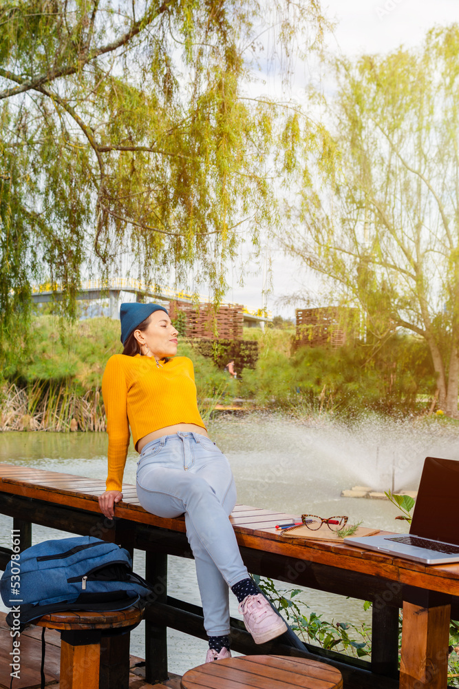 Attractive latin female student on college campus relaxing sitting on a wooden table while enjoying the sunset light