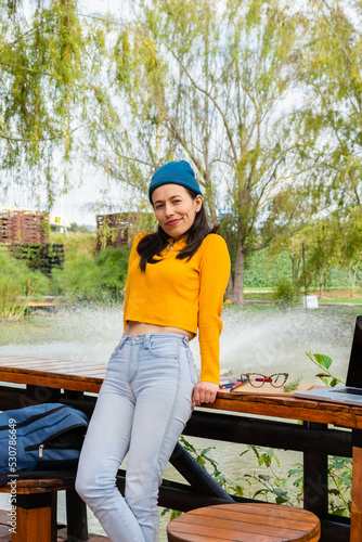 Attractive Latin student on campus resting leaning on a wooden table looking directly at the camera with her laptop next to her