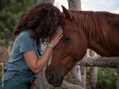 Close up of brunette woman forming bond with an anglo- arab horse behind a wood fence in a field.