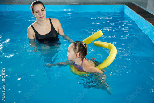 Preschool girllearning to swim in pool with foam noodle with young trainer photo