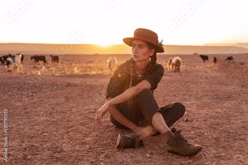 A girl in a hat sits on the sand in the desert at sunset. Goats grazing in the background. Morocco. © Nataliya