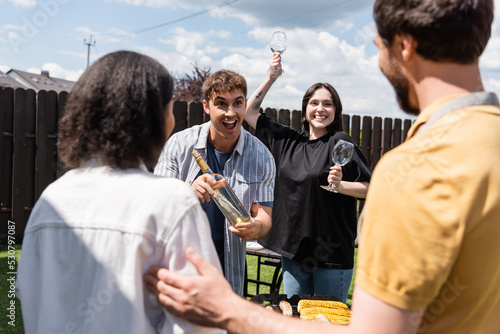 Cheerful man and woman holding wine near blurred multiethnic friends and food on bbq in backyard