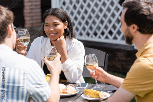 Smiling bi-racial woman holding glass of wine near blurred friends and food in backyard
