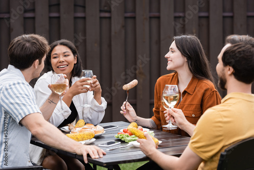 cheerful multiethnic friends holding glasses with wine and having meal during bbq party in backyard