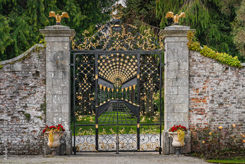 Beautiful, ornate wrought iron gate with golden details in Powerscourt gardens, Wicklow, Ireland. Brick wall with closed gate and forest in background photo