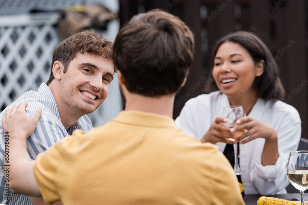 cheerful multiethnic friends smiling near blurred man on foreground during bbq party on backyard cheerful multiethnic friends smiling near blurred man on foreground during bbq party on backyard