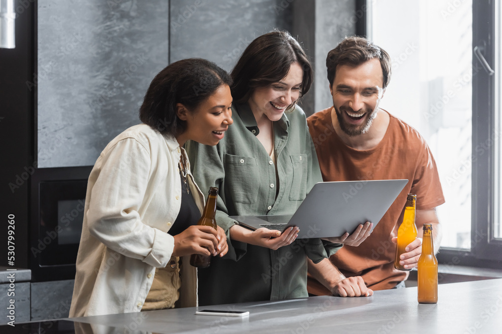 Obraz premium happy woman holding laptop near interracial friends with bottles of beer in kitchen