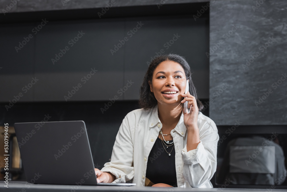 happy bi-racial woman talking on smartphone near laptop on kitchen worktop happy bi-racial woman talking on smartphone near laptop on kitchen worktop
