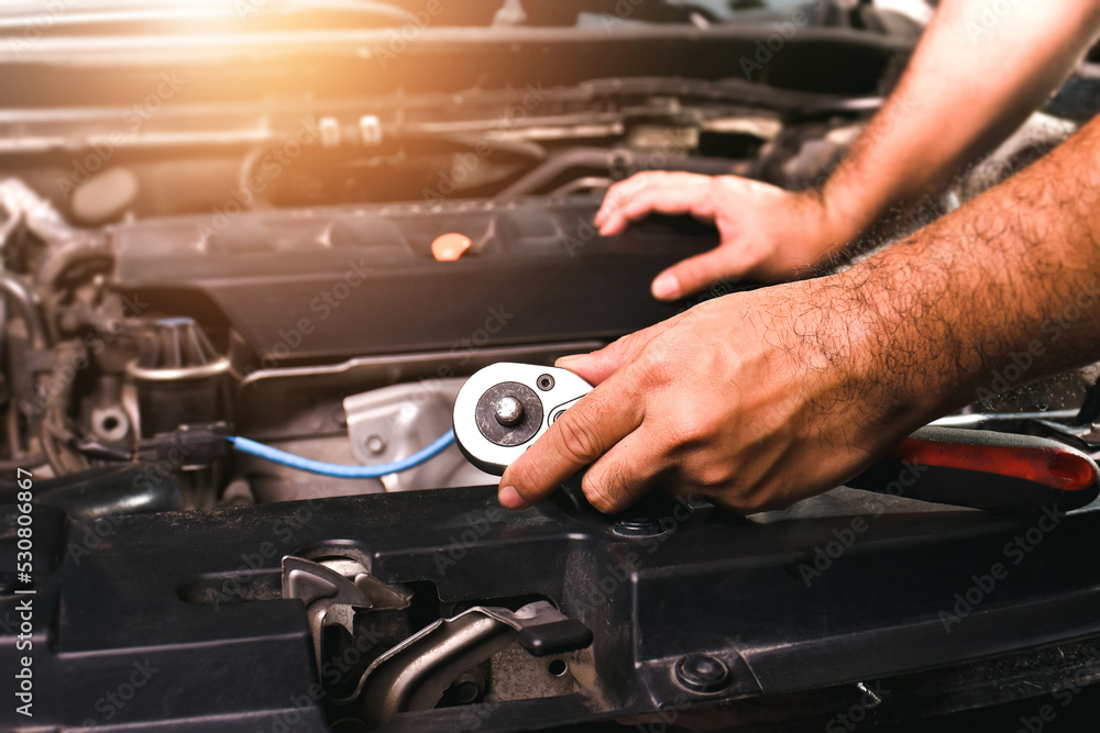 The mechanic is holding a metal socket wrench and putting his hand on the engine compartment of a car
