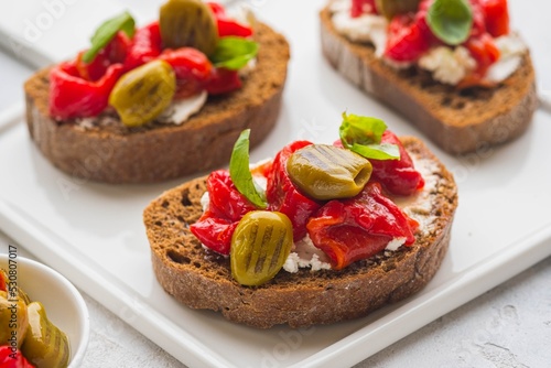 Open bruschetta sandwich with curd cheese, marinated roasted peppers and grilled olives on a white ceramic board on a light background. Sandwich recipes. Antipasti.