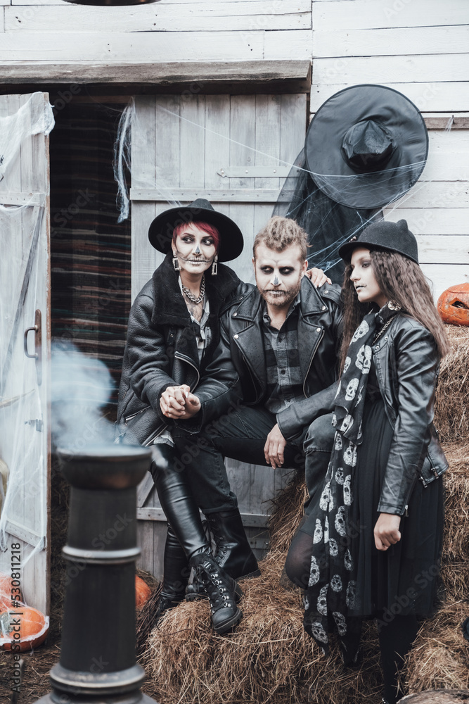 Scary family, mother, father, daughters celebrating halloween. Terrifying black skull half-face makeup and witch costumes, stylish images.Horror,fun at children's party in barn on street.Hats,jackets