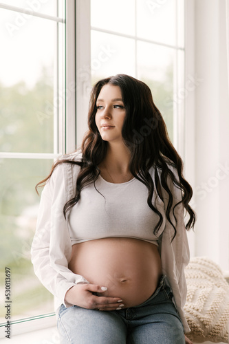 Beautiful pregnant girl with curls in a white shirt and jeans in a bright studio with a stylish interior, the concept of a happy pregnancy and family Free Download Beautiful pregnant girl with curls in a white shirt and jeans in a bright studio with a stylish interior, the concept of a happy pregnancy and family - GFXLoad
