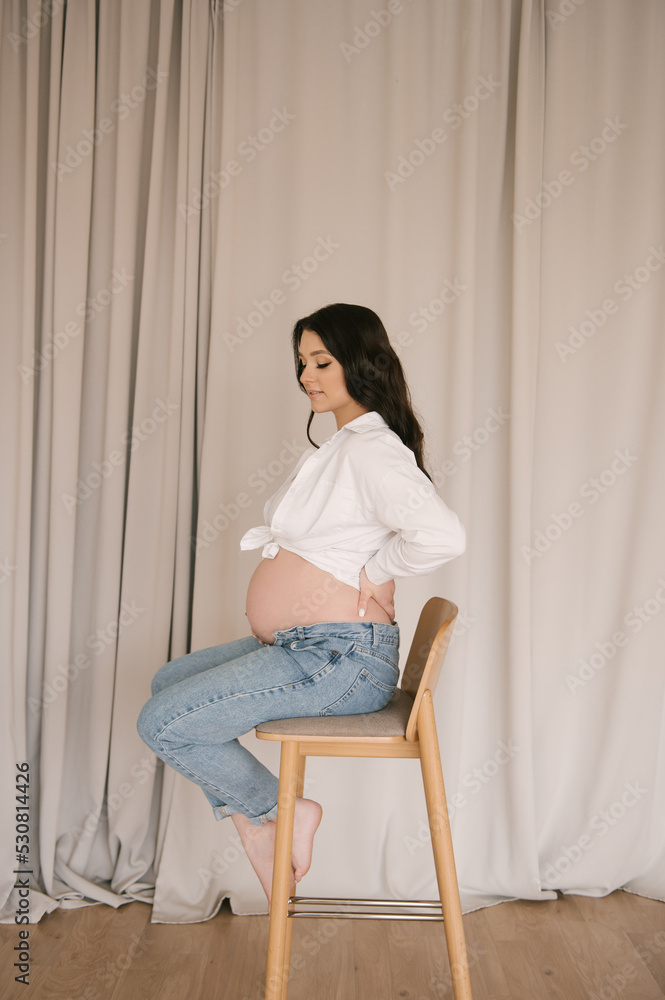 Beautiful pregnant girl with curls in a white shirt and jeans in a bright studio with a stylish interior, the concept of a happy pregnancy and family Beautiful pregnant girl with curls in a white shirt and jeans in a bright studio with a stylish interior, the concept of a happy pregnancy and family