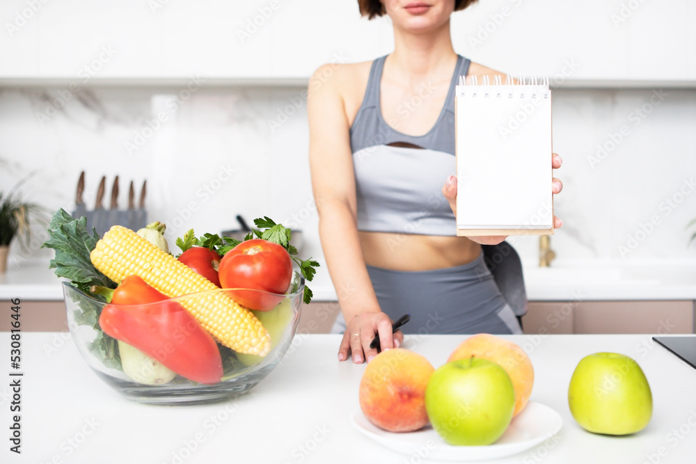 Close up photo of slender fit woman in sportswear demonstrating her diary sketchbook with empty space and many vegetables and fruits on the table Close up photo of slender fit woman in sportswear demonstrating her diary sketchbook with empty space and many vegetables and fruits on the table