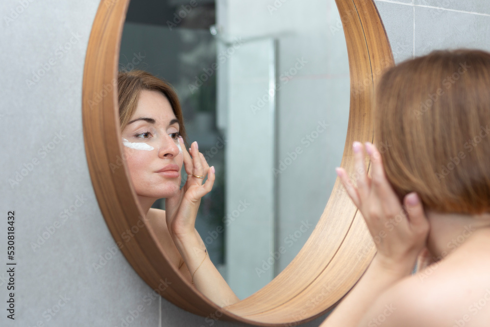Attractive woman looking at mirror and applying moisturizing cream on cheeks in bathroom. Groomed young girl doing skincare morning routine after taking a shower Attractive woman looking at mirror and applying moisturizing cream on cheeks in bathroom. Groomed young girl doing skincare morning routine after taking a shower