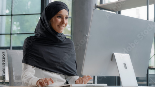 Leinwand Poster Young arab businesswoman in hijab working on computer smiling enjoying office wo