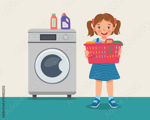 happy little girl holding basket full of clothes doing laundry daily chores with washing machine 
