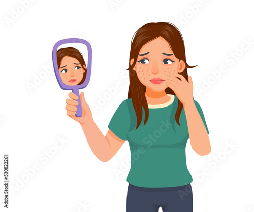 Anxious young woman holding mirror looking at acne spot pimples on her face