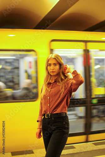 a blonde girl in the subway is standing in casual clothes near a passing yellow subway 