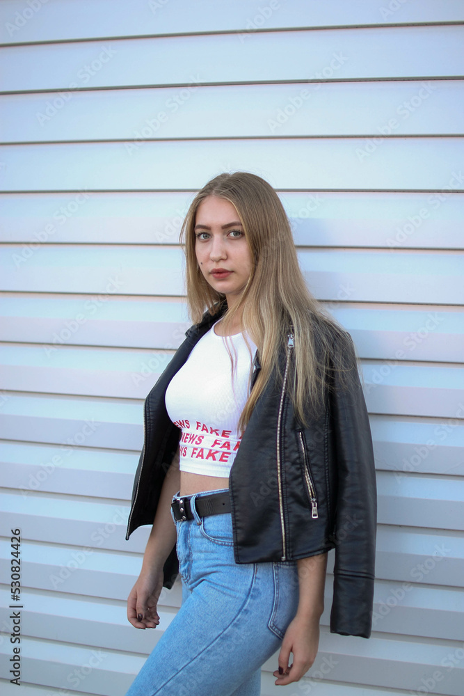 a blonde girl in a leather jacket and light jeans stands near a plain white wall and crosses her arms , portrait photo a blonde girl in a leather jacket and light jeans stands near a plain white wall and crosses her arms , portrait photo