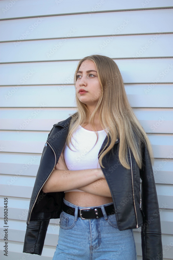 a blonde girl in a leather jacket and light jeans stands near a plain white wall and crosses her arms , portrait photo a blonde girl in a leather jacket and light jeans stands near a plain white wall and crosses her arms , portrait photo