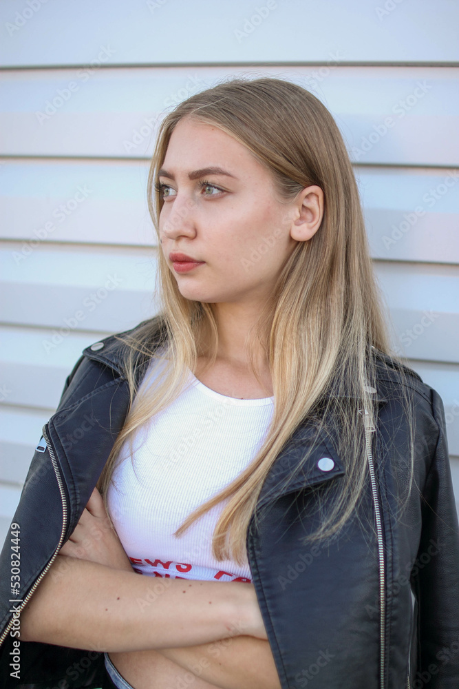 a blonde girl in a leather jacket and light jeans stands near a plain white wall and crosses her arms , portrait photo
