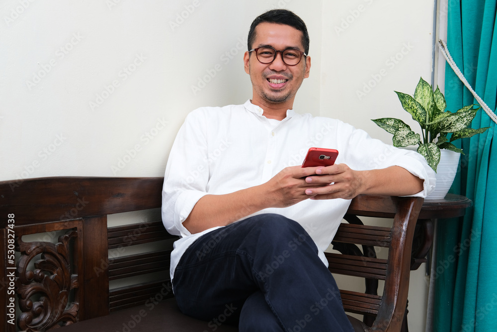 Adult Asian man sitting in a couch smiling when holding mobile phone