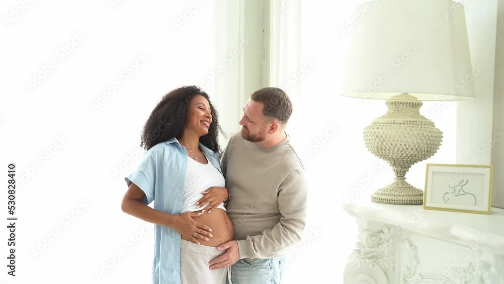 White man touching belly of his pregnant black wife. Pregnant woman and loving husband hugging tummy at home. Feeling baby movement by multiethnic couple on pregnant belly. White man touching belly of his pregnant black wife. Pregnant woman and loving husband hugging tummy at home. Feeling baby movement by multiethnic couple on pregnant belly.