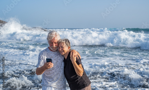 Happy caucasian senior couple standing on the beach at sunset looking at mobile phone - smiling pensioners enjoying free time, sea vacation or retirement #530831876