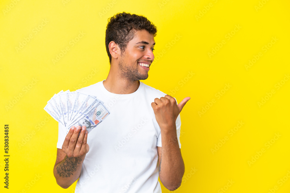 Young brazilian man taking a lot of money isolated on yellow background pointing to the side to present a product Young brazilian man taking a lot of money isolated on yellow background pointing to the side to present a product