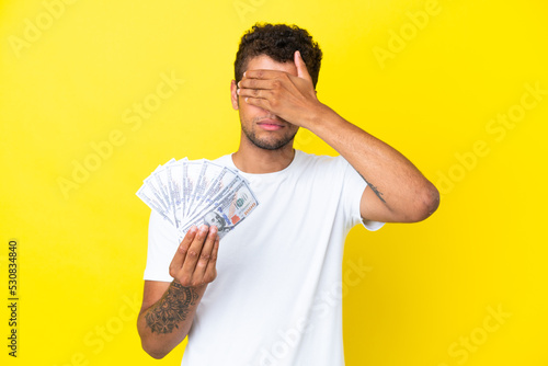 Young brazilian man taking a lot of money isolated on yellow background covering eyes by hands. Do not want to see something
