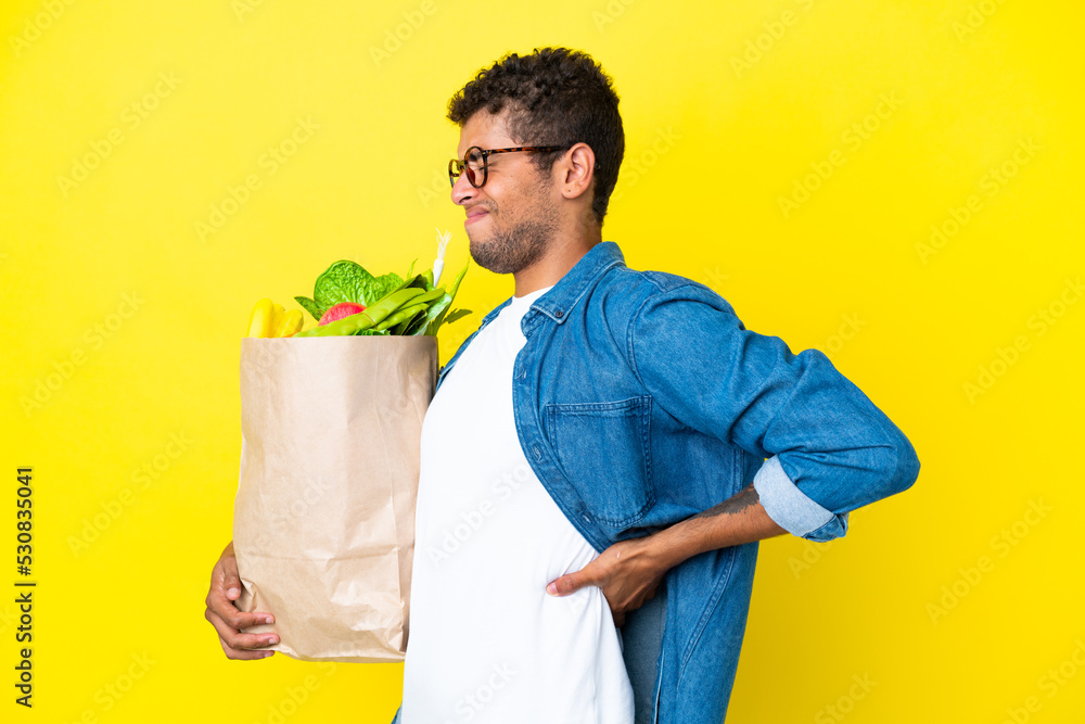 Young Brazilian man holding a grocery shopping bag isolated on yellow background suffering from backache for having made an effort Young Brazilian man holding a grocery shopping bag isolated on yellow background suffering from backache for having made an effort