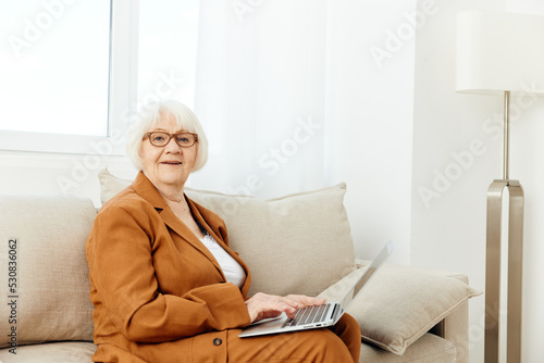 a pleasant, sweetly smiling elderly old lady is sitting with a laptop on her lap on a beige sofa dressed in a stylish brown suit and looking at the camera © Tatiana