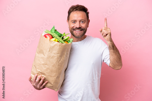 Middle age man holding a grocery shopping bag isolated on pink background pointing up a great idea