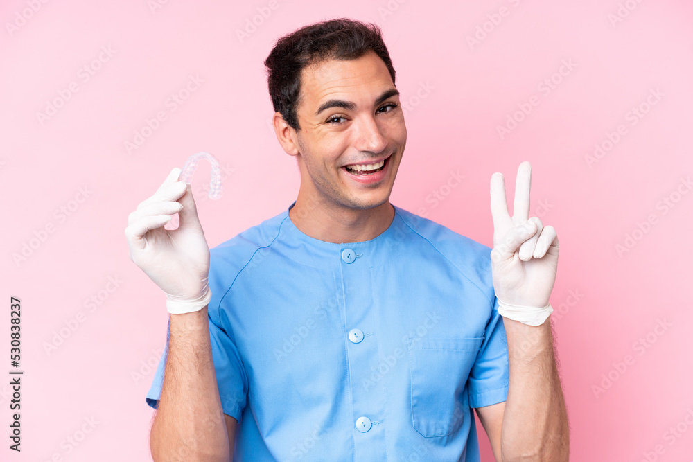 Dentist caucasian man holding invisible braces isolated on pink background smiling and showing victory sign Dentist caucasian man holding invisible braces isolated on pink background smiling and showing victory sign