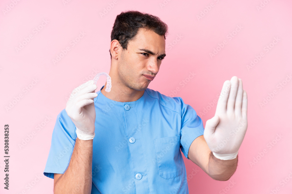 Dentist caucasian man holding invisible braces isolated on pink background making stop gesture and disappointed Dentist caucasian man holding invisible braces isolated on pink background making stop gesture and disappointed