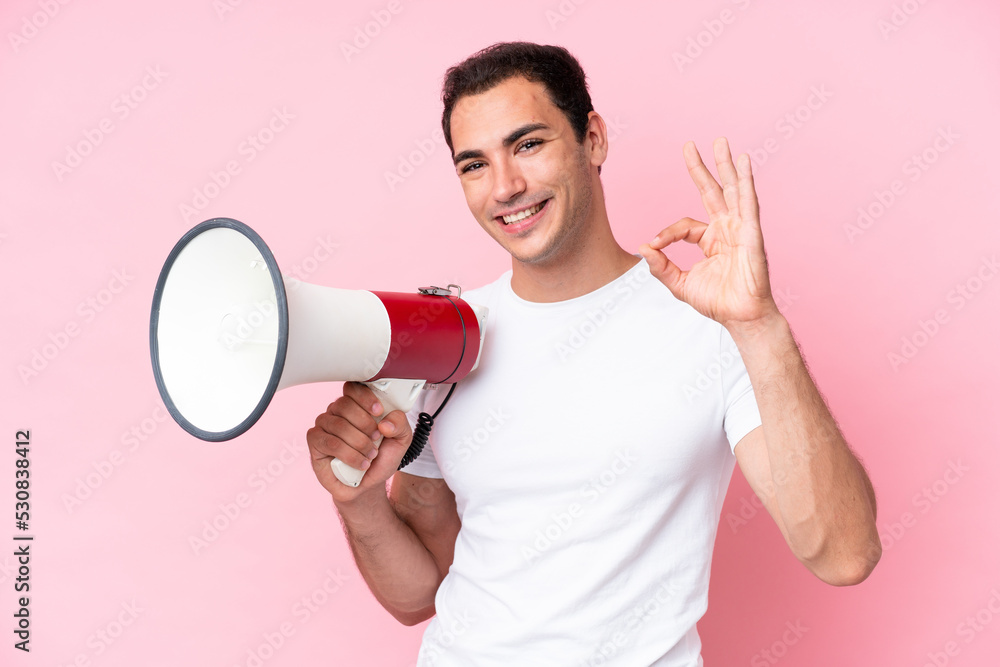 Young caucasian man isolated on pink background holding a megaphone and showing ok sign with fingers Young caucasian man isolated on pink background holding a megaphone and showing ok sign with fingers
