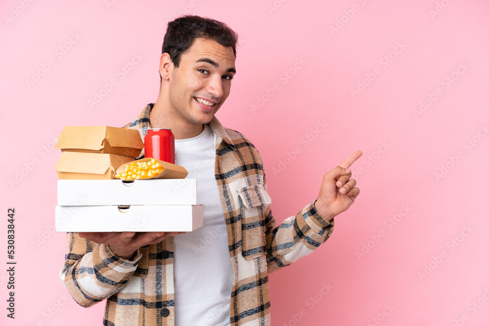 Young caucasian man holding fast food isolated on pink background pointing finger to the side Young caucasian man holding fast food isolated on pink background pointing finger to the side