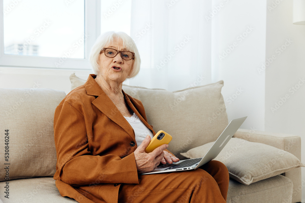 an elderly businesswoman is sitting at home in a bright apartment on a beige sofa dressed in a stylish brown suit with a laptop and a smartphone in her hands looking funny into the camera an elderly businesswoman is sitting at home in a bright apartment on a beige sofa dressed in a stylish brown suit with a laptop and a smartphone in her hands looking funny into the camera