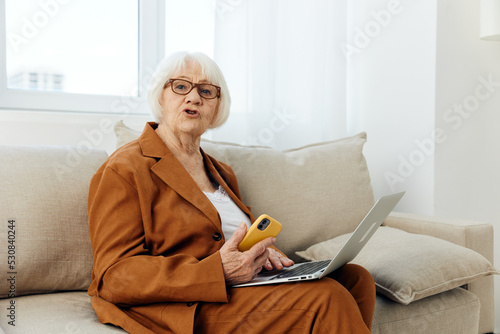 an elderly businesswoman is sitting at home in a bright apartment on a beige sofa dressed in a stylish brown suit with a laptop and a smartphone in her hands looking funny into the camera © Tatiana